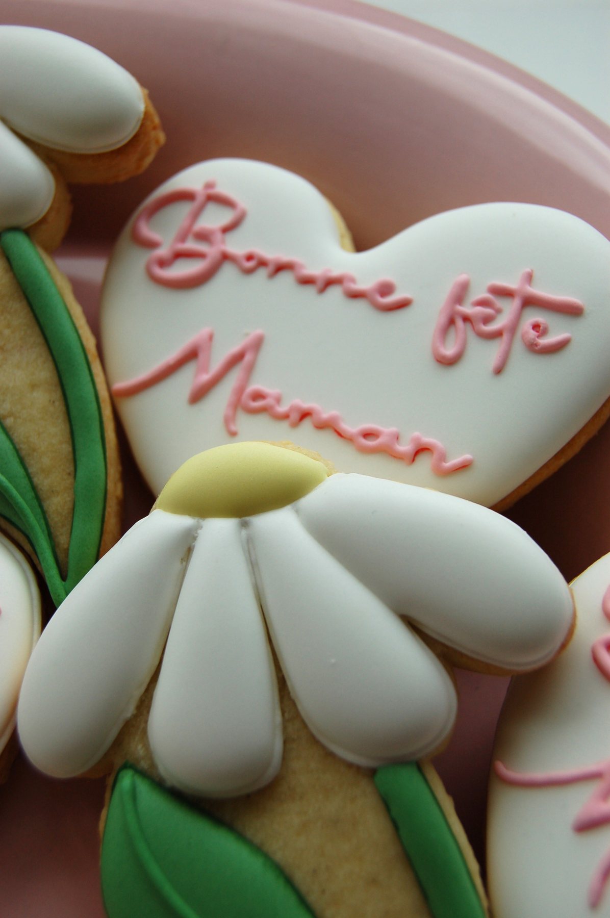 Biscuit cœur blanc calligraphié Bonne fête Maman, posé à côté d'une marguerite